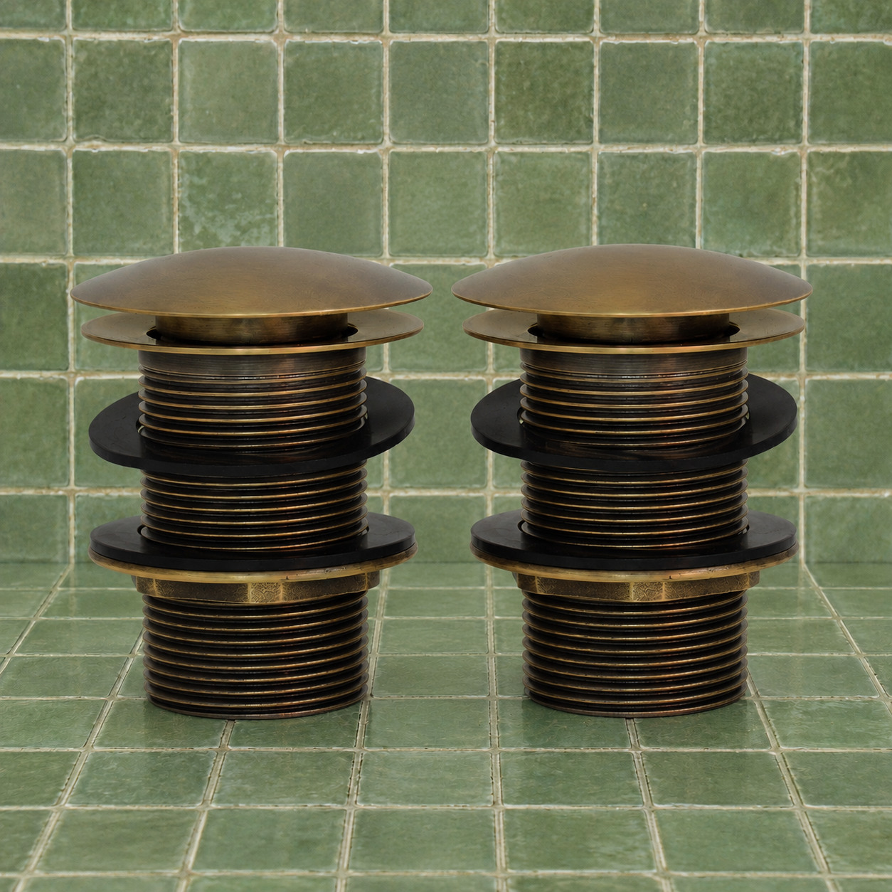 A bronze-colored metal plumbing fixture component, likely a drain stopper or valve assembly, shown against a background of green square ceramic tiles.