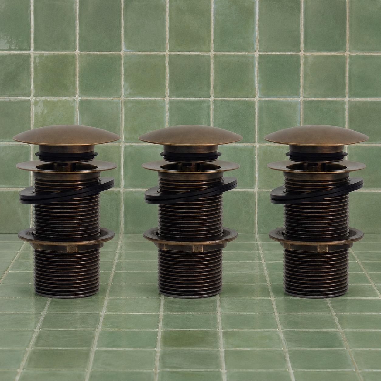 A bronze-colored metal plumbing fixture component, likely a drain stopper or valve assembly, shown against a background of green square ceramic tiles.