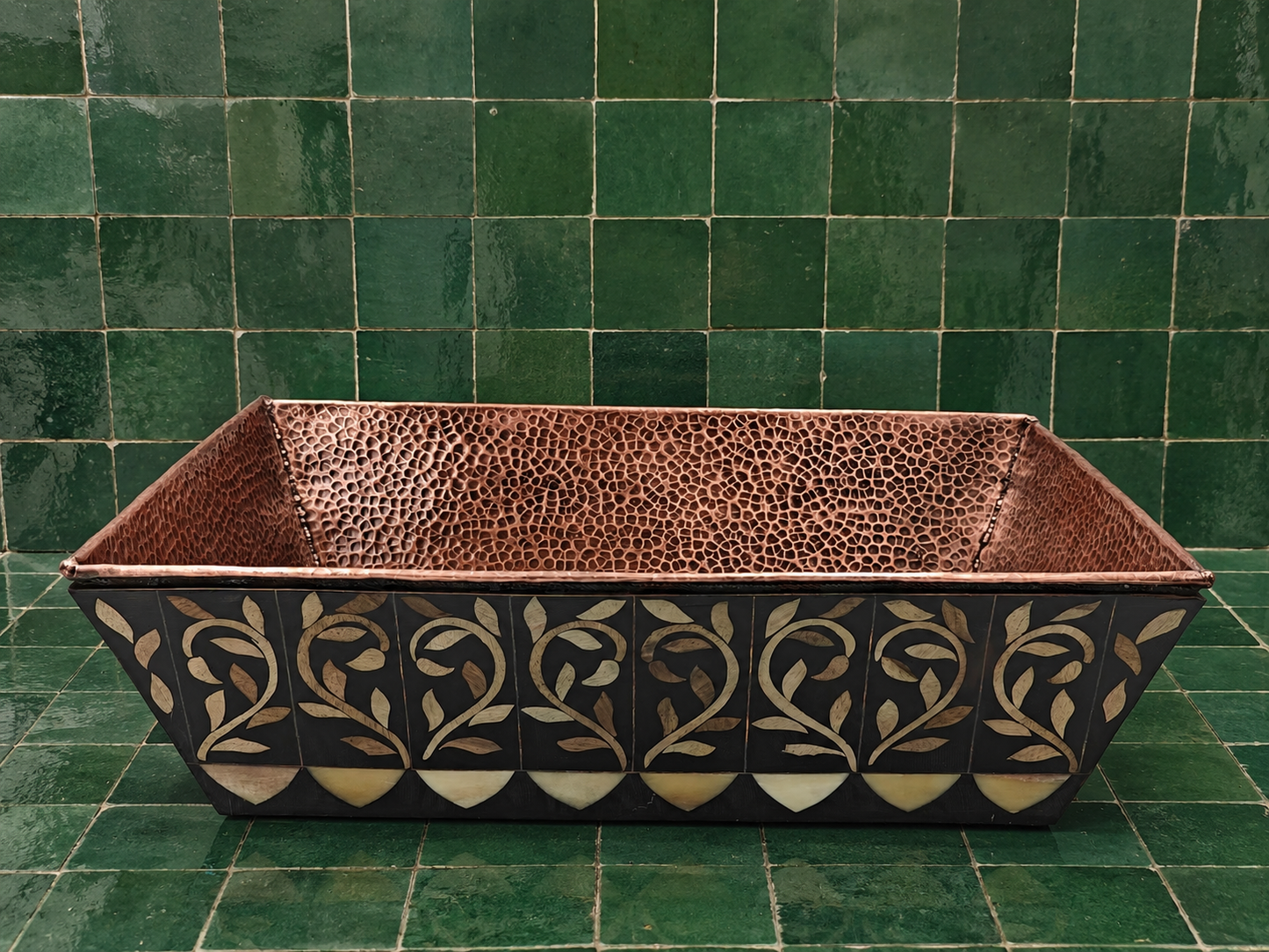 A rectangular copper planter with a textured interior and a black exterior decorated with gold and beige floral inlay, set against a green tiled background.