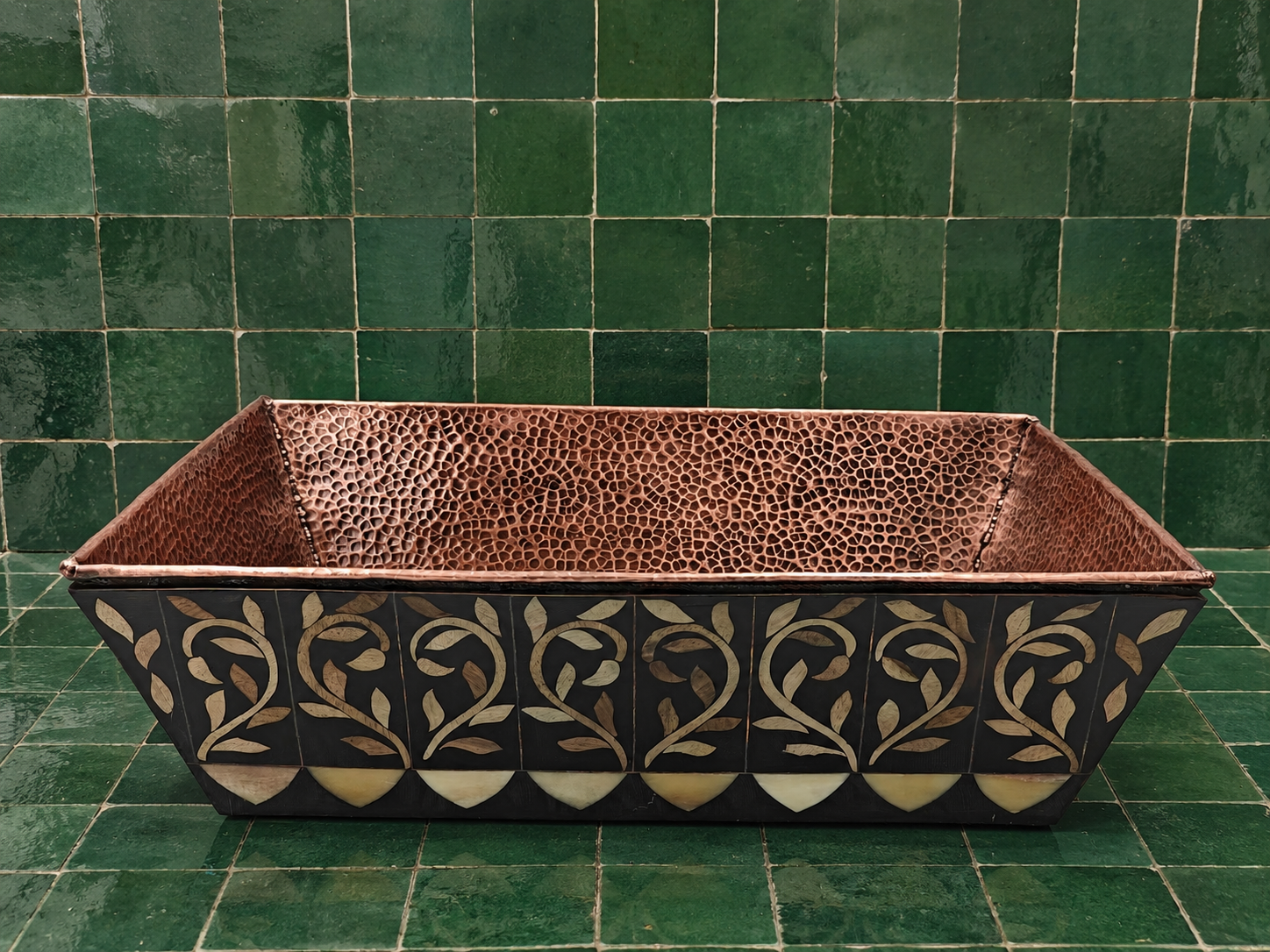 A rectangular copper planter with a textured interior and a black exterior decorated with gold and beige floral inlay, set against a green tiled background.