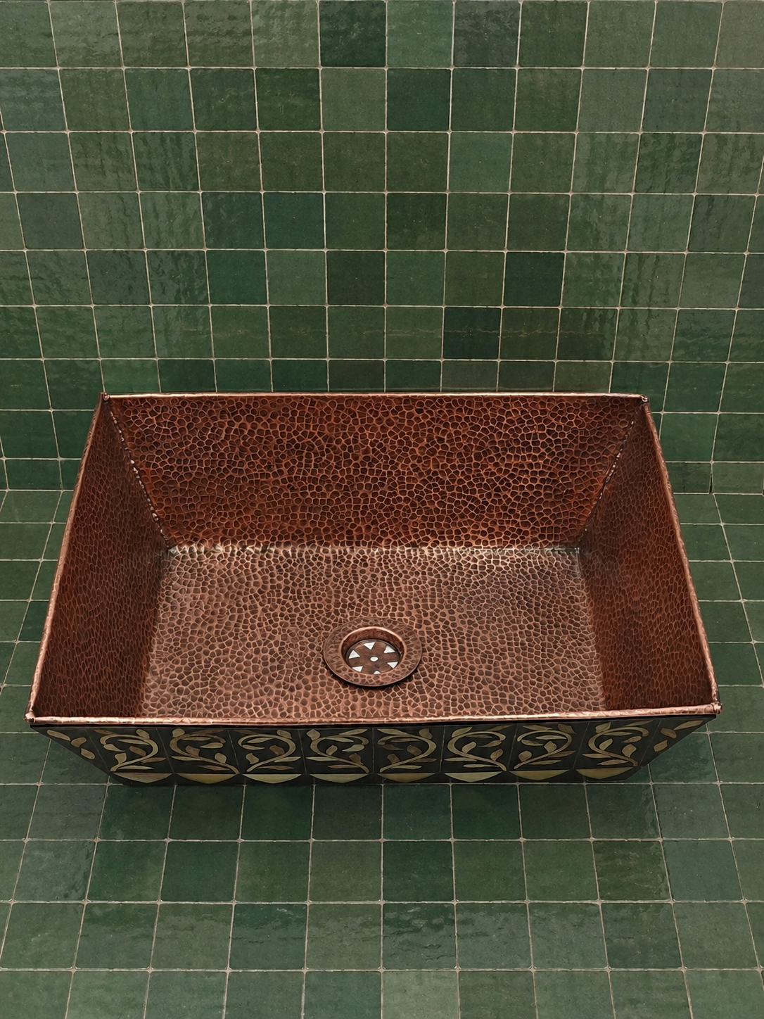 A rectangular hammered copper bathroom sink with a decorative dark patterned apron, shown against a green tiled wall and floor.