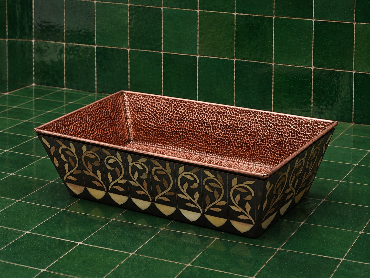 A rectangular hammered copper planter with a black and gold floral patterned base, set against a green tiled background.