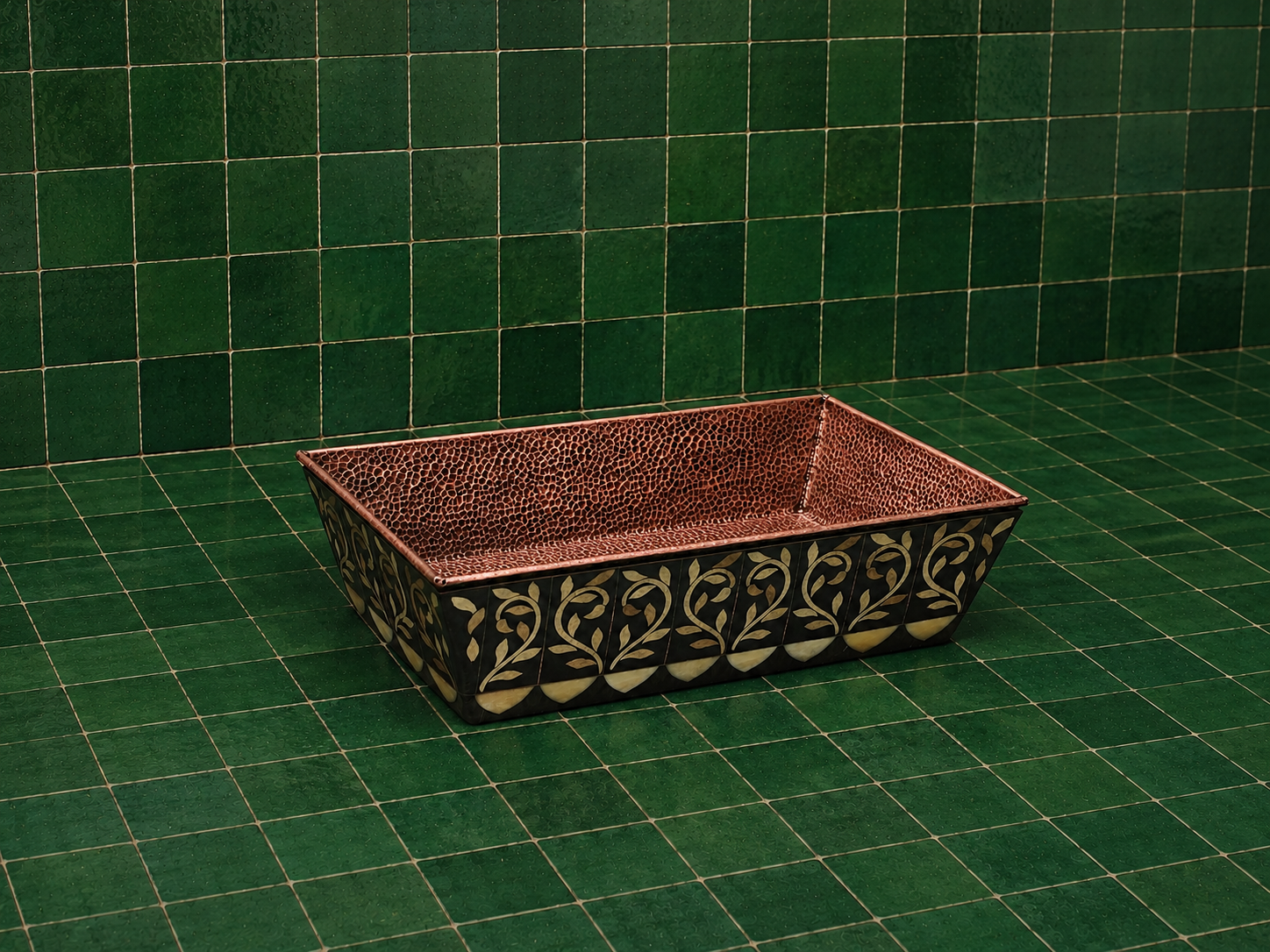 A rectangular hammered copper sink basin with a black and gold floral patterned lower half, set against a green tiled background.