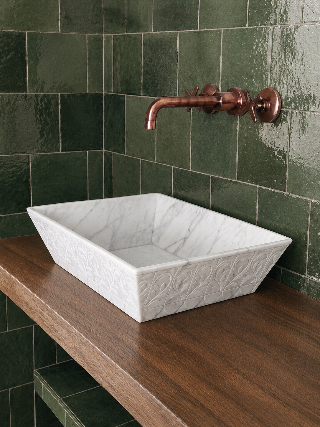 A rectangular white marble vessel sink with embossed leaf patterns, set on a dark wood countertop against a glossy dark green tiled wall with a copper faucet.