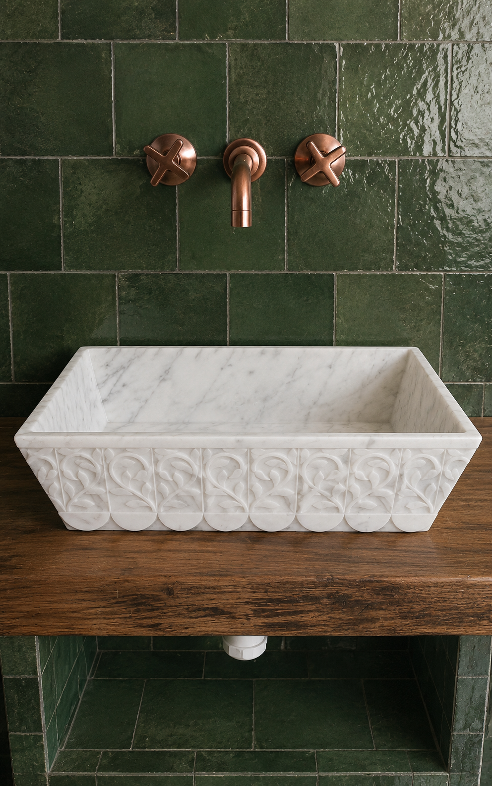 A rectangular white marble vessel sink with an embossed leaf pattern, mounted on a rustic dark wood vanity against a glossy dark green tiled wall with a copper faucet.