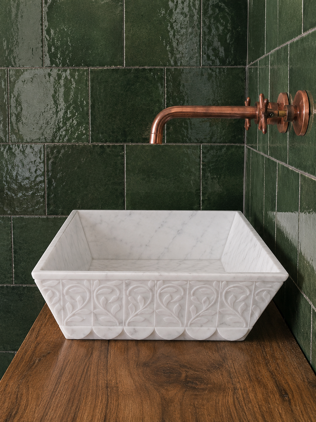 A rectangular white marble vessel sink with embossed scrollwork, set on a dark wood countertop against a glossy dark green tiled wall with a copper faucet.