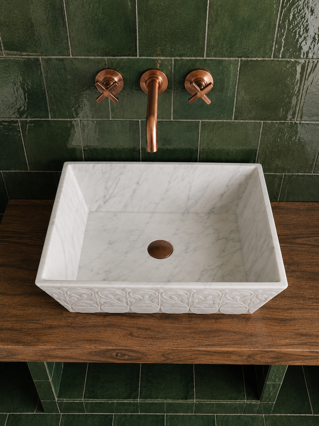 A rectangular white marble vessel sink with embossed detailing on the front, mounted on a dark wood vanity top against a glossy dark green tiled wall. A copper-finished wall-mounted faucet and two cross-handle valves are visible above the sink.
