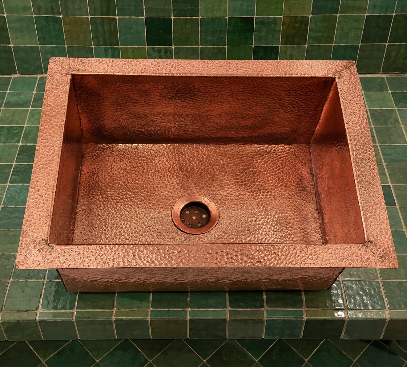 A hammered copper farmhouse sink with a square basin and integrated drain, set into a green tiled countertop.