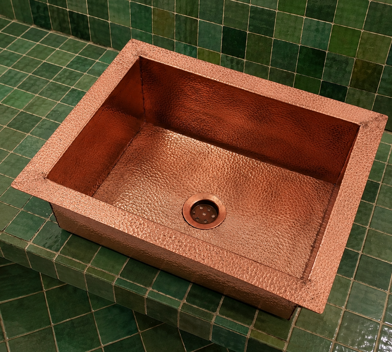 A rectangular hammered copper farmhouse sink with a matching drain, set into a green tiled countertop and backsplash.