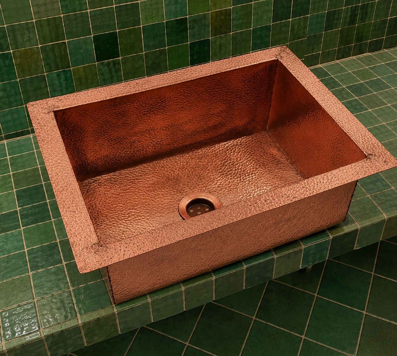 A hammered copper farmhouse sink with a square drop-in design, shown installed on a green tiled countertop with matching green wall tiles.