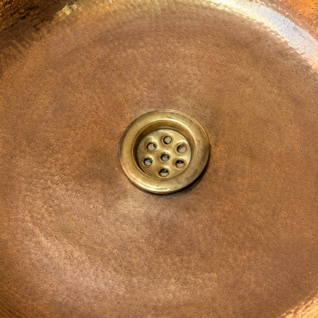 Close-up of a hammered copper sink basin with a round brass drain in the center.