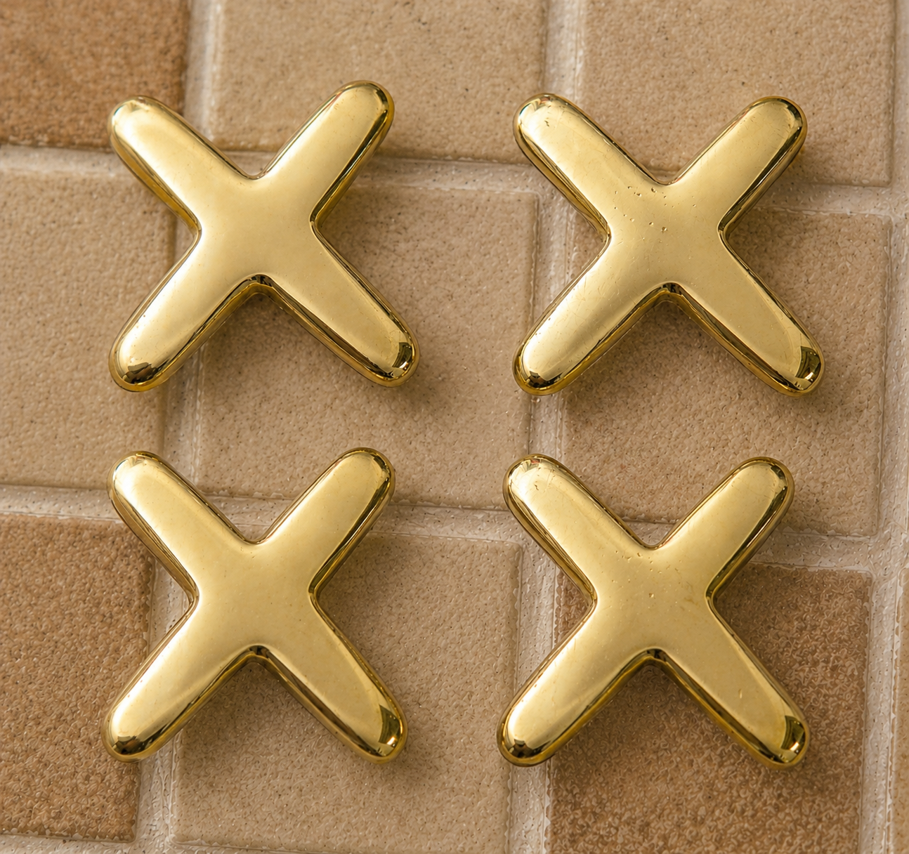 Four shiny gold X-shaped earrings arranged in a 2x2 grid on a beige tiled background.