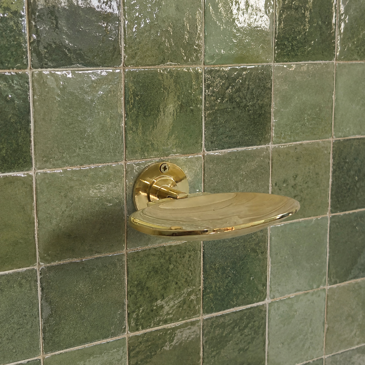 A polished brass wall-mounted soap dish with a circular backplate, mounted on a green glazed tile wall.