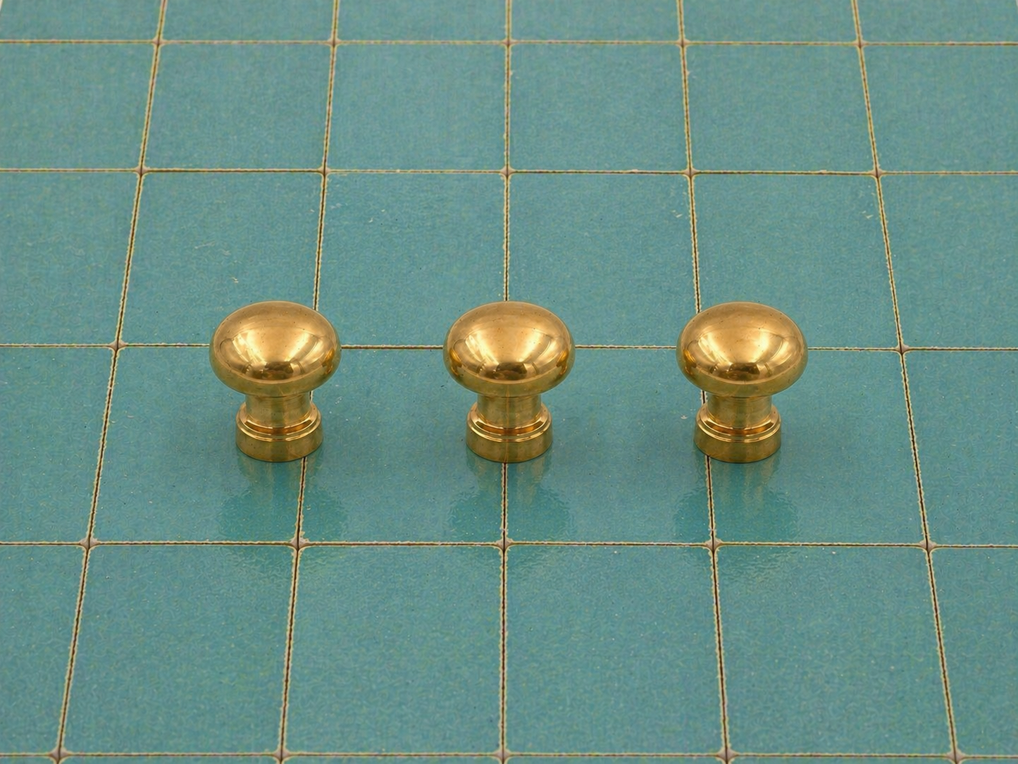 Three small round brass cabinet knobs arranged in a row on a teal grid cutting mat.