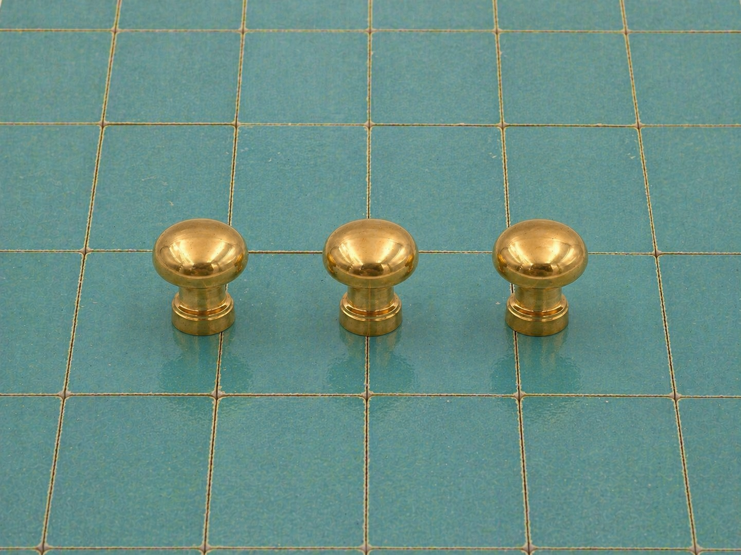 Three small round brass cabinet knobs arranged in a row on a teal grid cutting mat.