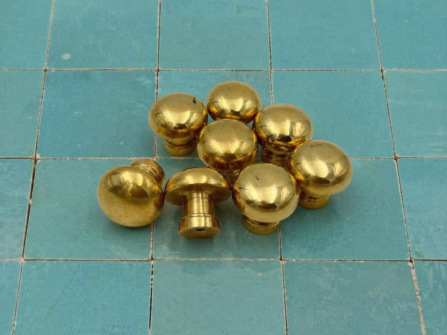 A group of shiny brass cabinet knobs, including round ball knobs and mushroom-shaped pull knobs, arranged on a blue-green tiled surface.