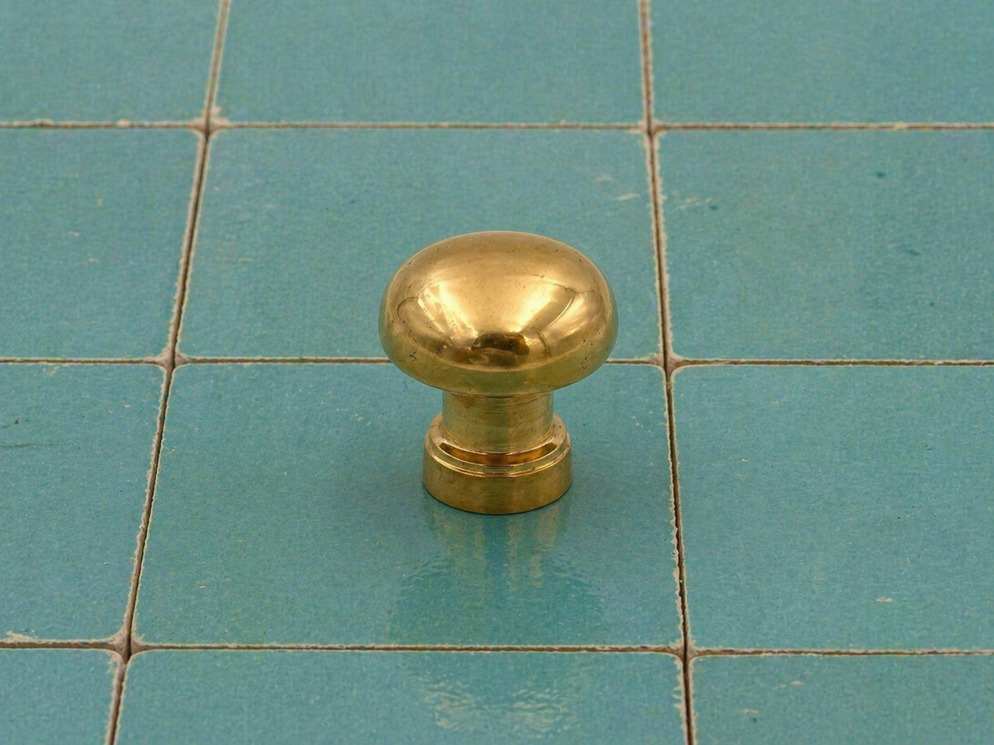 A round, polished brass cabinet knob standing upright on a teal tiled surface.
