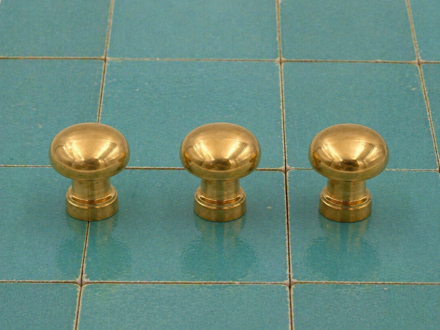 Three round brass cabinet knobs arranged in a row on a teal tiled surface.