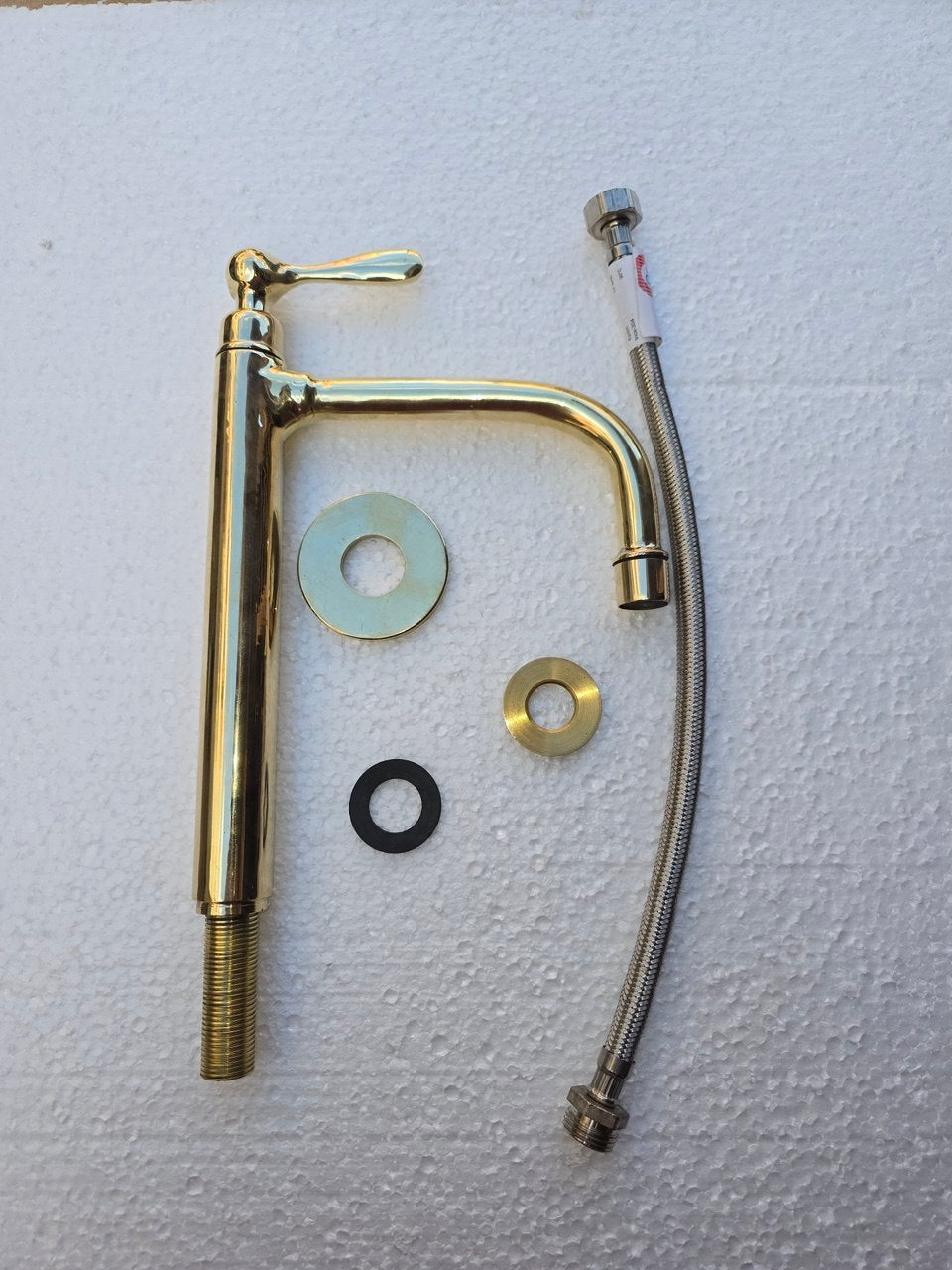 A brass-finished single-handle faucet with a curved spout, shown with a braided metal supply hose, mounting washers, and a black O-ring on a white textured background.