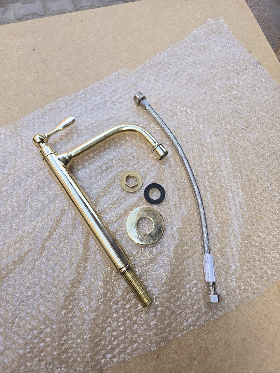 A brass wall-mounted kitchen faucet with a curved gooseneck spout and lever handle, shown with a braided metal supply line and mounting hardware on bubble wrap over a cardboard surface.