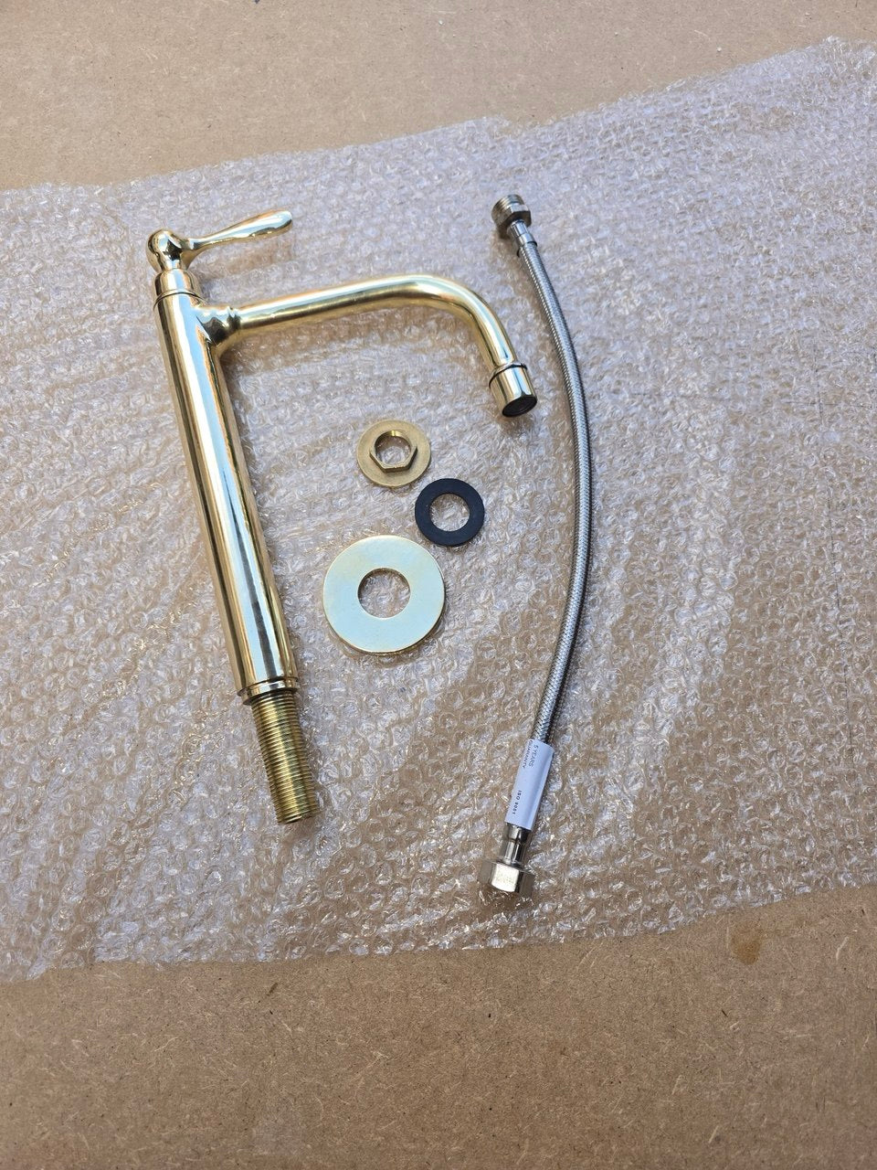 A metal faucet conversion kit with a curved brass spout, threaded pipe section, flexible metal hose, and mounting washers, laid out on bubble wrap over a brown cardboard surface.