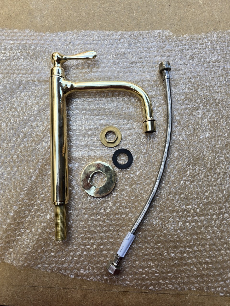 A brass wall-mounted faucet with a curved spout and lever handle, shown with mounting hardware and a braided metal supply line on bubble wrap.